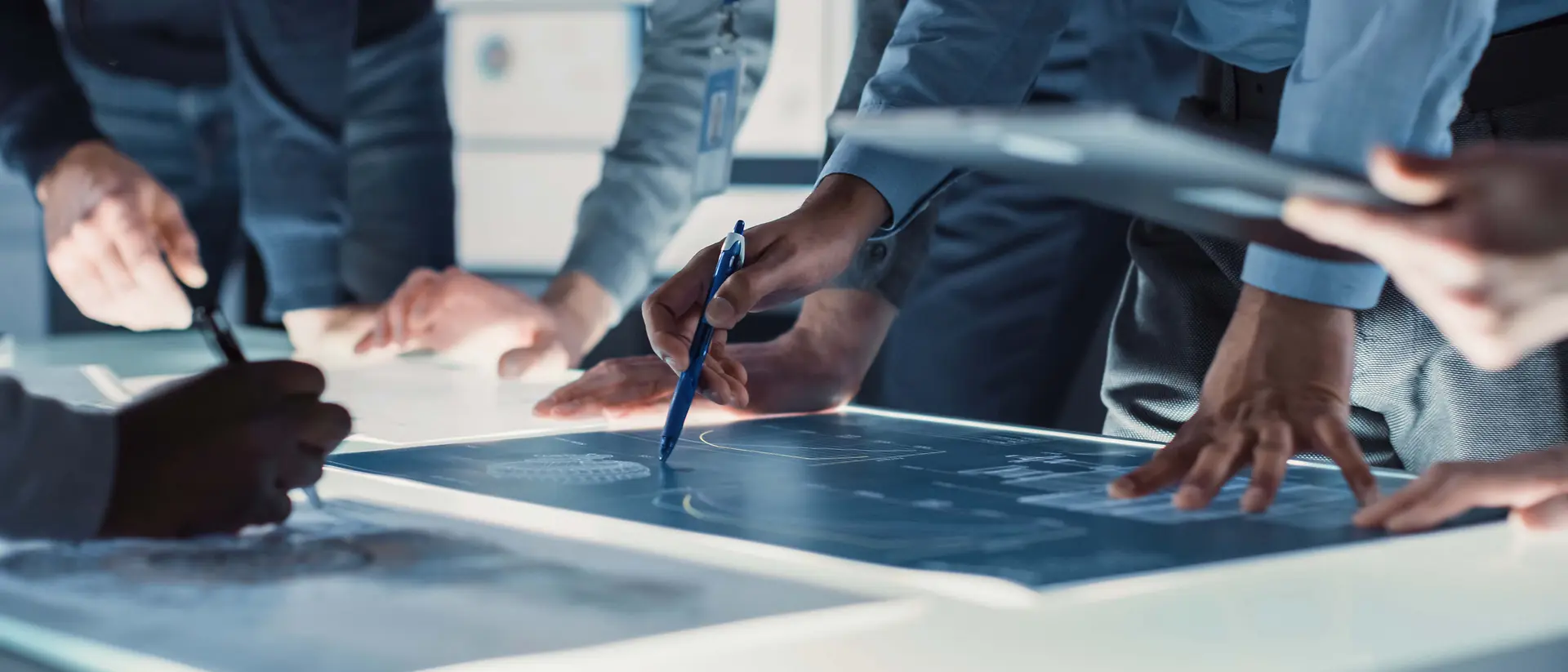 Close-up view of several people gathered around a large illuminated table, reviewing and marking technical blueprints with pens and digital devices.