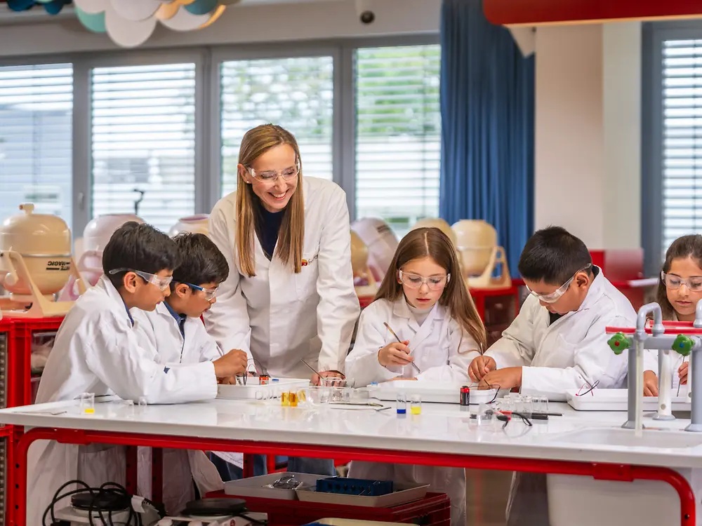 A group of children wearing lab coats work together on science experiments at a classroom table, while an Simone Bagel-Trah guides them. The room is equipped with scientific materials, bright lighting, and organized shelves.