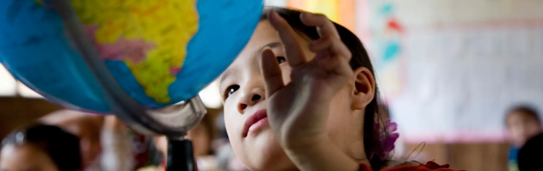 A child in a classroom reaches toward a large globe positioned on a desk, with colorful maps and posters visible on the walls in the background.