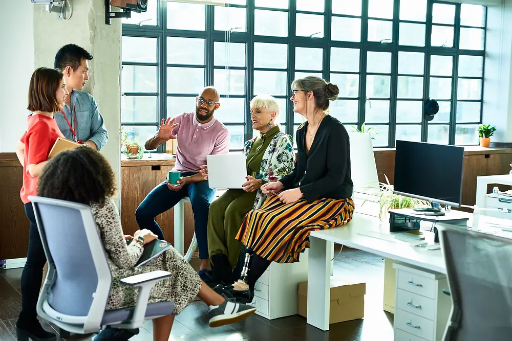 A small group of individuals sitting and standing in a bright, open office, talking together near desks and large windows.