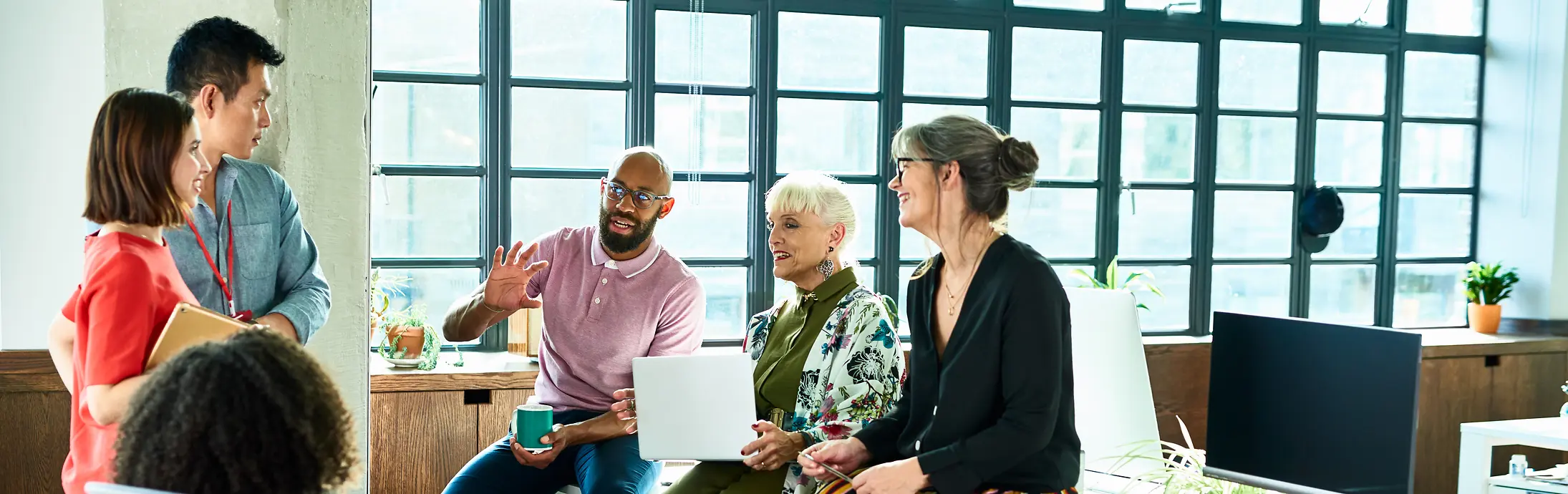 A small group of individuals sitting and standing in a bright, open office, talking together near desks and large windows.