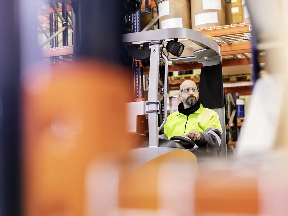 
A warehouse worker wearing a high‑visibility jacket operates a forklift, surrounded by shelves stocked with large containers and equipment