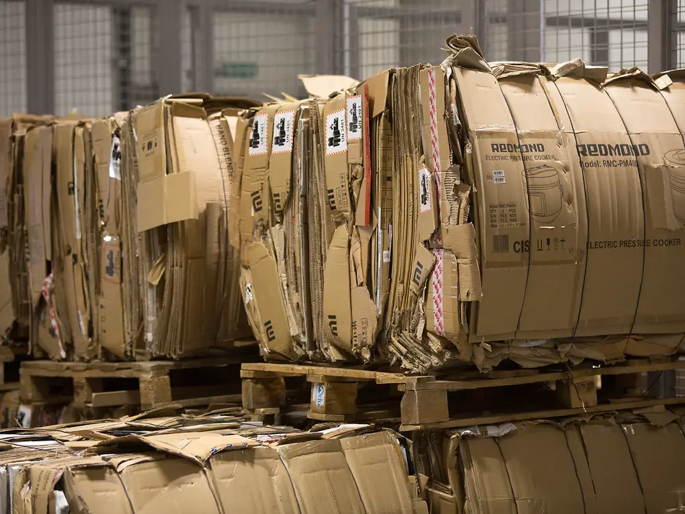 Stacks of flattened, bundled cardboard boxes arranged on wooden pallets in an industrial warehouse setting, ready for recycling or disposal.