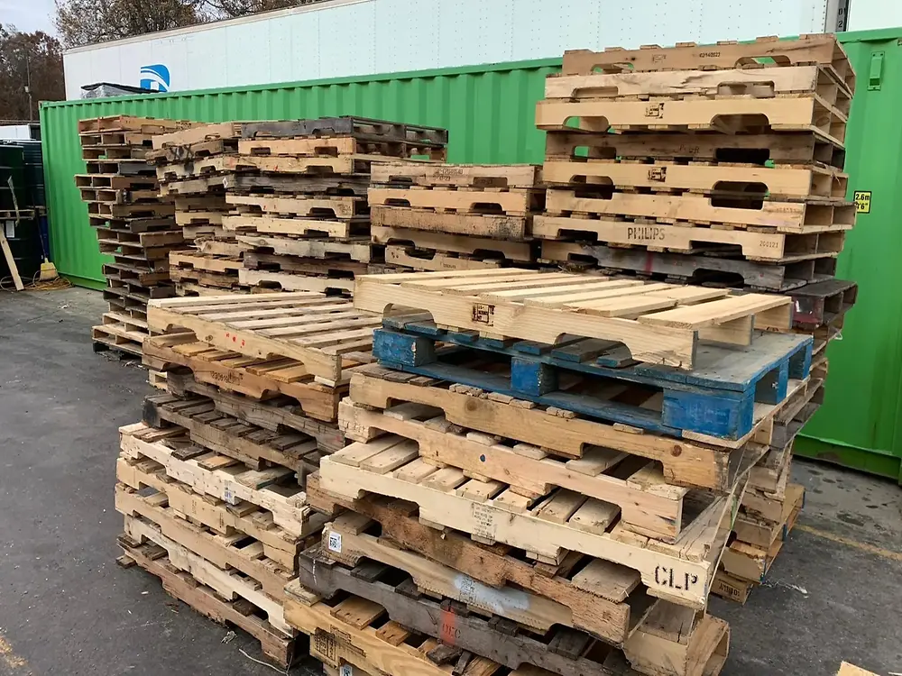  Large stacks of wooden pallets piled outdoors on an asphalt surface, with a green shipping container and other storage units in the background.