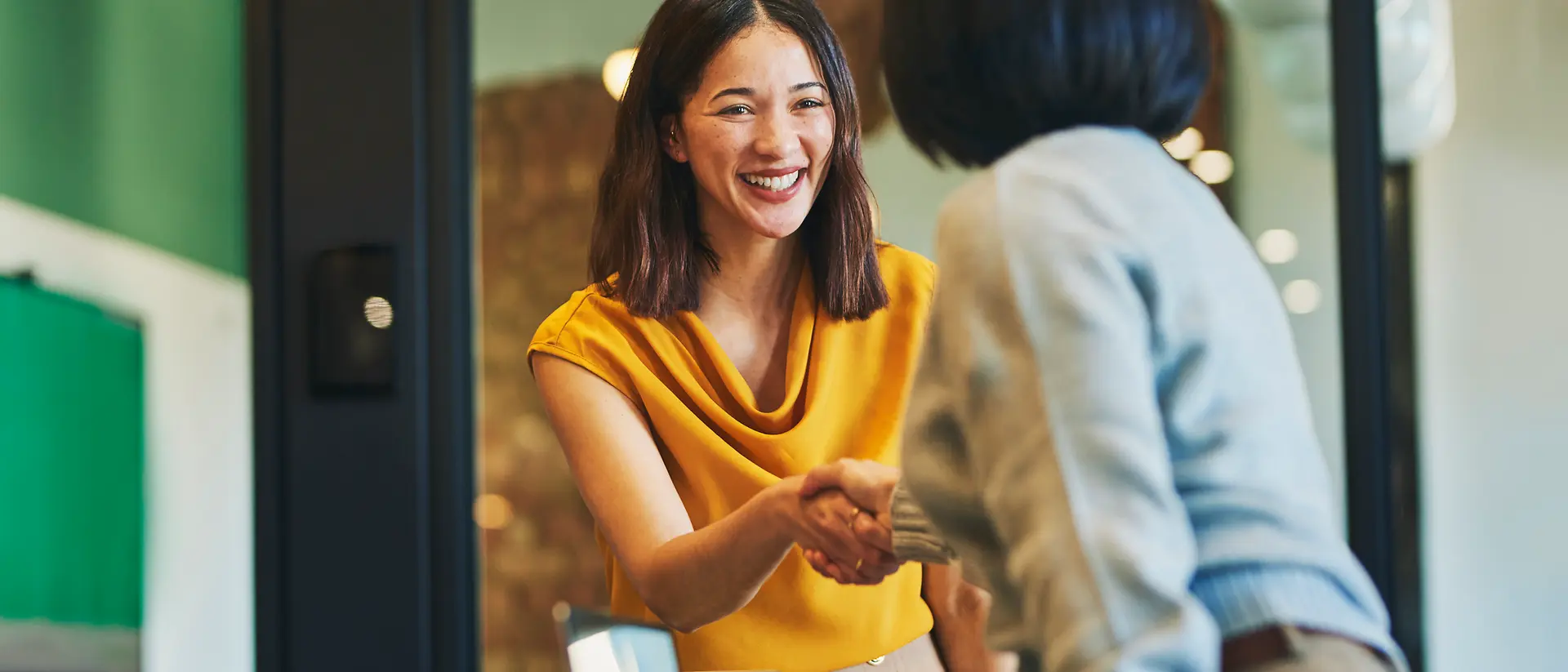 Zwei Personen geben sich über einen Tisch hinweg die Hand in einem modernen Besprechungsraum, im Vordergrund steht ein Laptop.