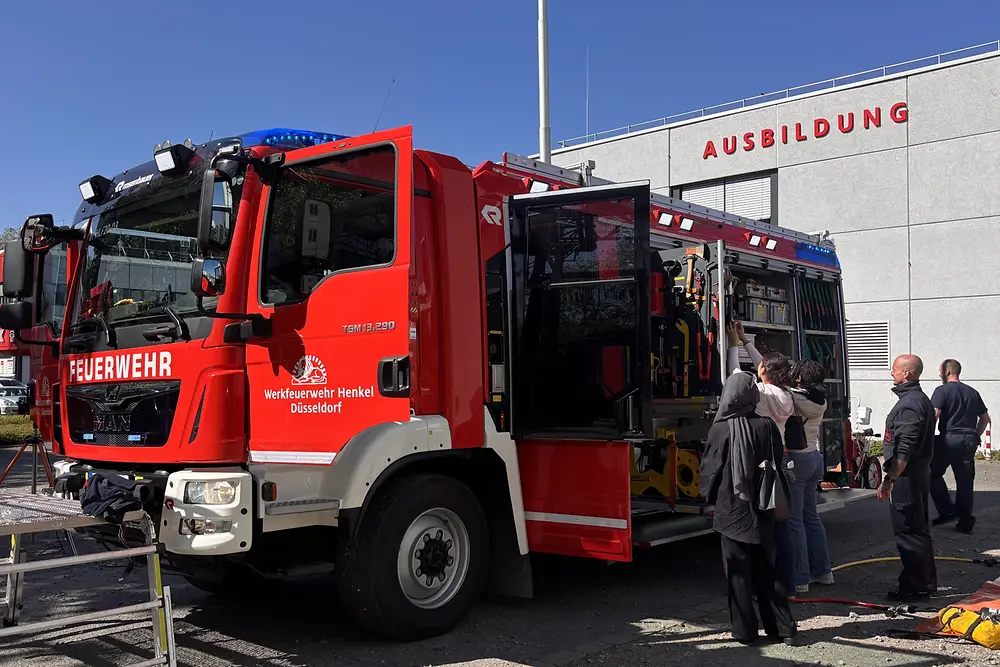 Ein Feuerwehrfahrzeug der Werkfeuerwehr Henkel steht mit geöffneten Geräteraumklappen vor dem Ausbildungsgebäude; drei Mädchen schauen sich die Ausrüstung an.