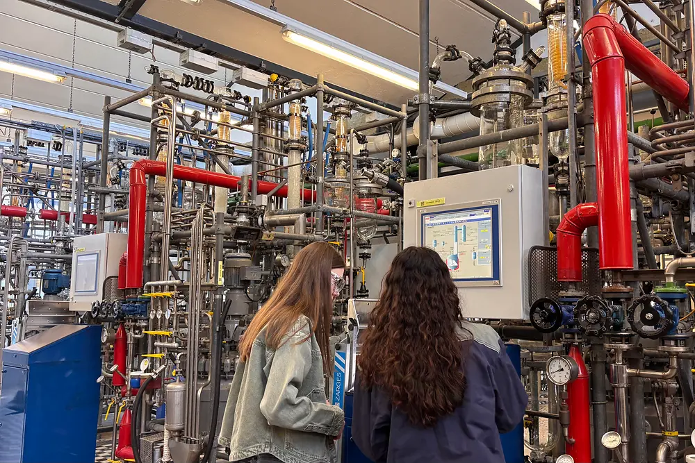 Zwei Mädchen stehen vor einer großen verfahrenstechnischen Anlage mit Rohrleitungen, Ventilen und digitalem Bedienpanel.