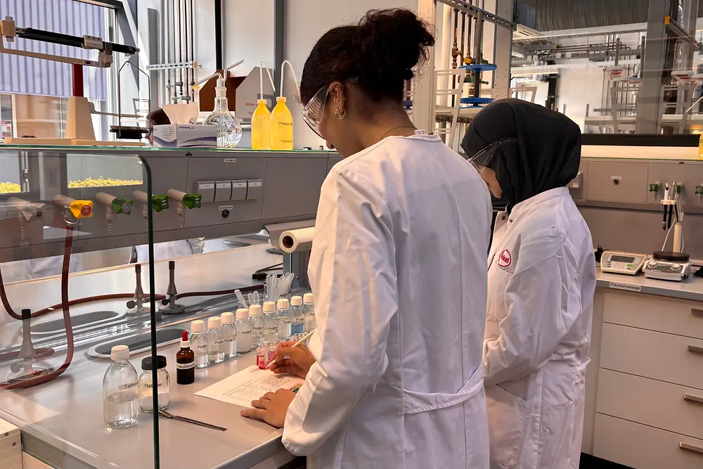 Zwei Mädchen im Labor führen einen Versuch durch; Reagenzflaschen, Messgeräte und Laborunterlagen stehen auf der Arbeitsfläche.
