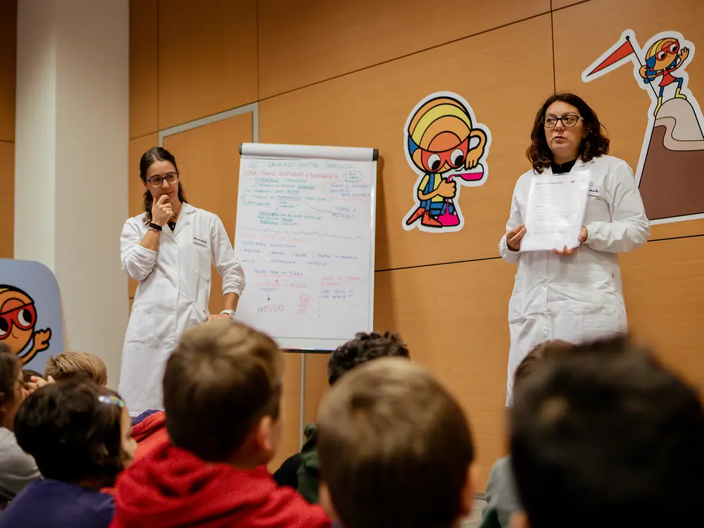Zwei Frauen in Laborkitteln stehen vor einer Gruppe von Kindern. Eine Person präsentiert ein Blatt Papier, die andere steht neben einem Flipchart mit Notizen. Der Raum ist mit Forscherwelt‑Illustrationen dekoriert.