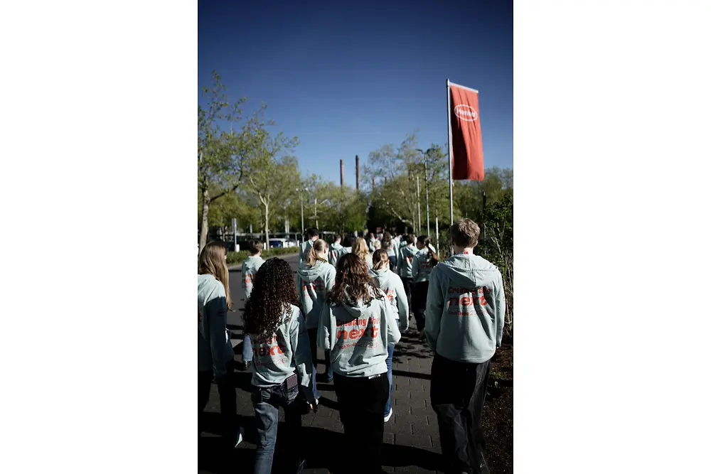 Einige Jugendliche gehen über das Henkel-Werkgelände; im Hintergrund sieht man Produktionsgebäude und eine rote Henkel-Fahne. 