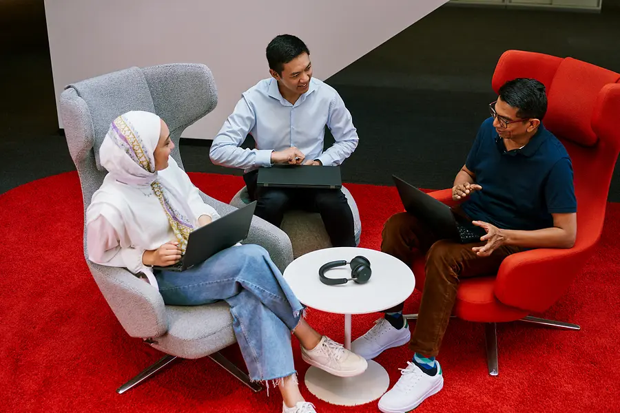Drei Kolleg:innen sitzen mit Laptops in einem modernen Office-Lounge-Bereich und tauschen sich aus. Sie sitzen in farbigen Sesseln um einen kleinen Tisch – typisch für ein kollaboratives Arbeitsmeeting.