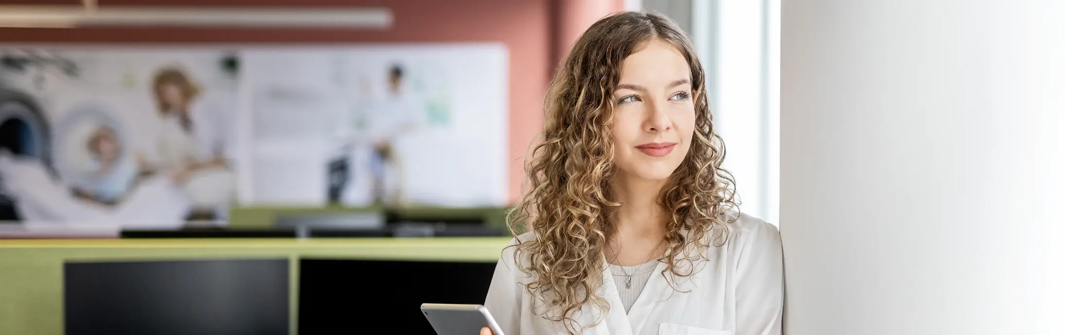 Eine junge Frau lehnt mit einem Laptop in der Hand an einer Wand in einem modernen Büro.