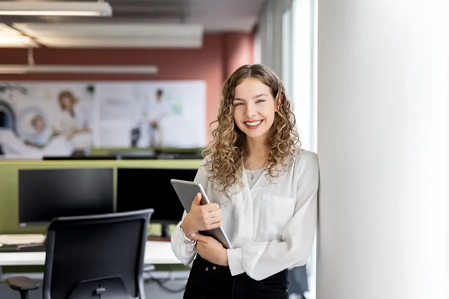 Eine junge Frau lehnt mit einem Laptop in der Hand an einer Wand in einem modernen Büro