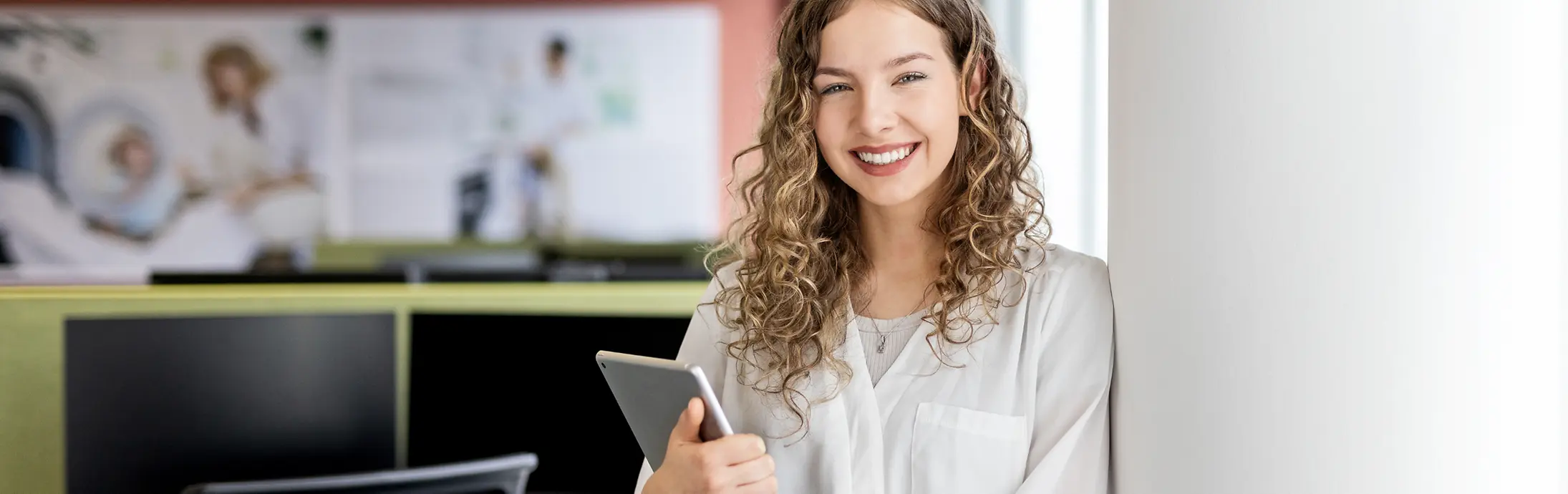 Eine junge Frau lehnt mit einem Laptop in der Hand an einer Wand in einem modernen Büro