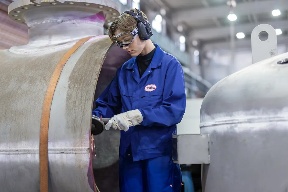 Ein Mitarbeitender mit Schutzbrille, Gehörschutz, Handschuhen und blauer Arbeitsjacke bearbeitet die Oberfläche eines großen Edelstahlbehälters in einer Produktionshalle