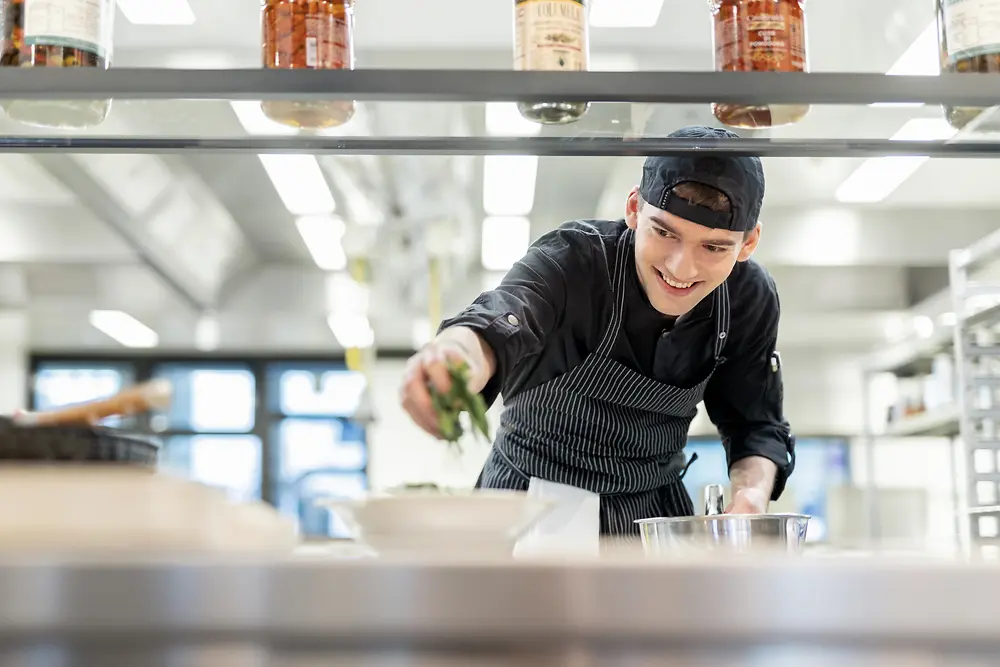 Ein Koch in einer professionellen Küche garniert ein Gericht mit frischen Kräutern. Im Hintergrund sind Edelstahlflächen und Küchenequipment sichtbar