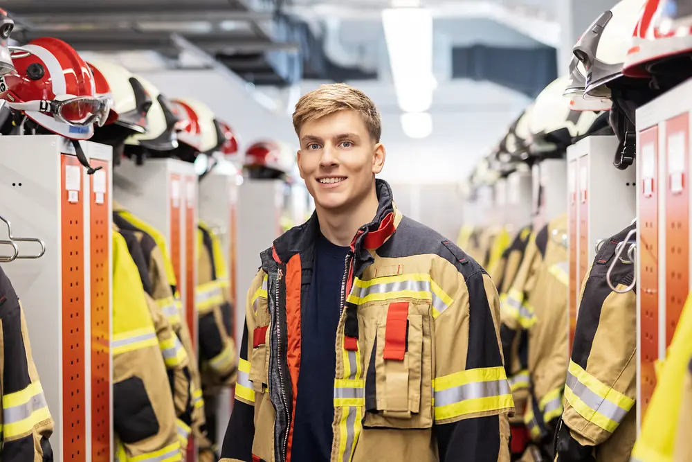 Ein Feuerwehrmitglied in vollständiger Schutzausrüstung steht in einer Feuerwache und hält Helm sowie Handschuhe in den Händen. Im Hintergrund sind Einsatzfahrzeuge zu sehen