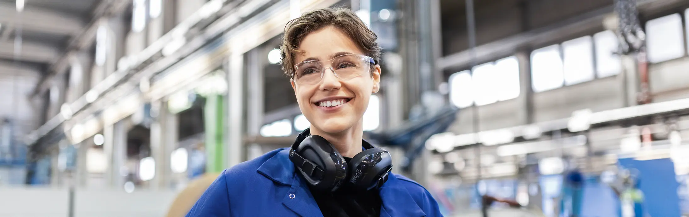 Ein junger Kollege steht mit Schutzbrille und blauem Arbeitskragen in einer Produktionsumgebung
