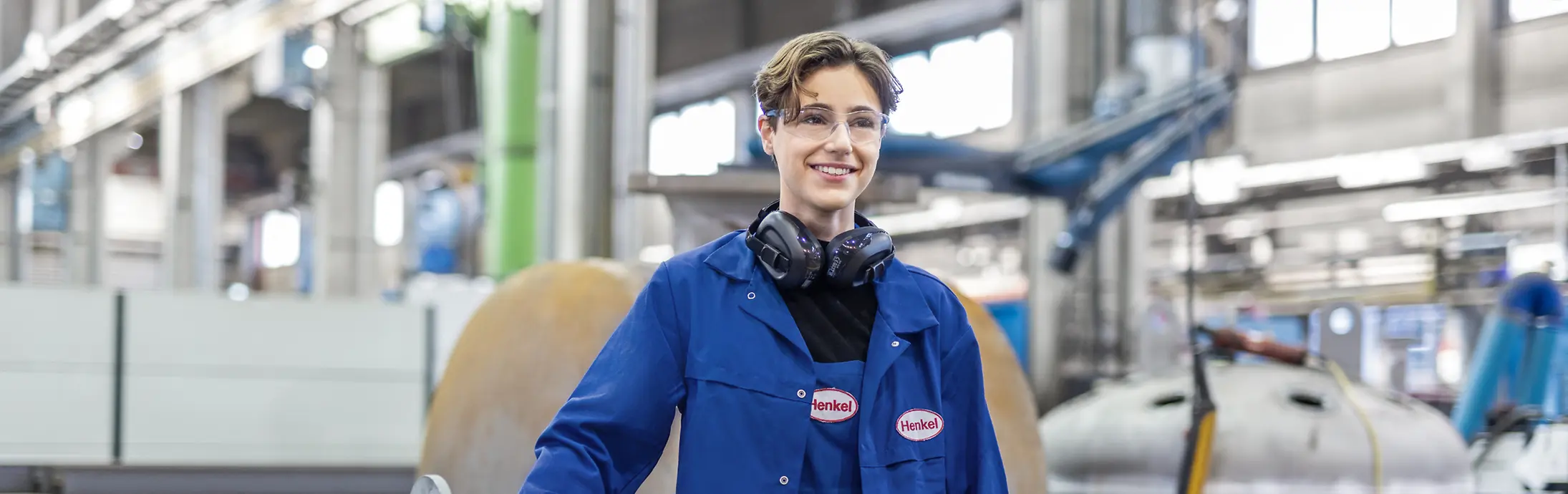 Eine junge Mitarbeiterin mit Schutzbrille und blauer Arbeitskleidung an einem Produktionsstandort