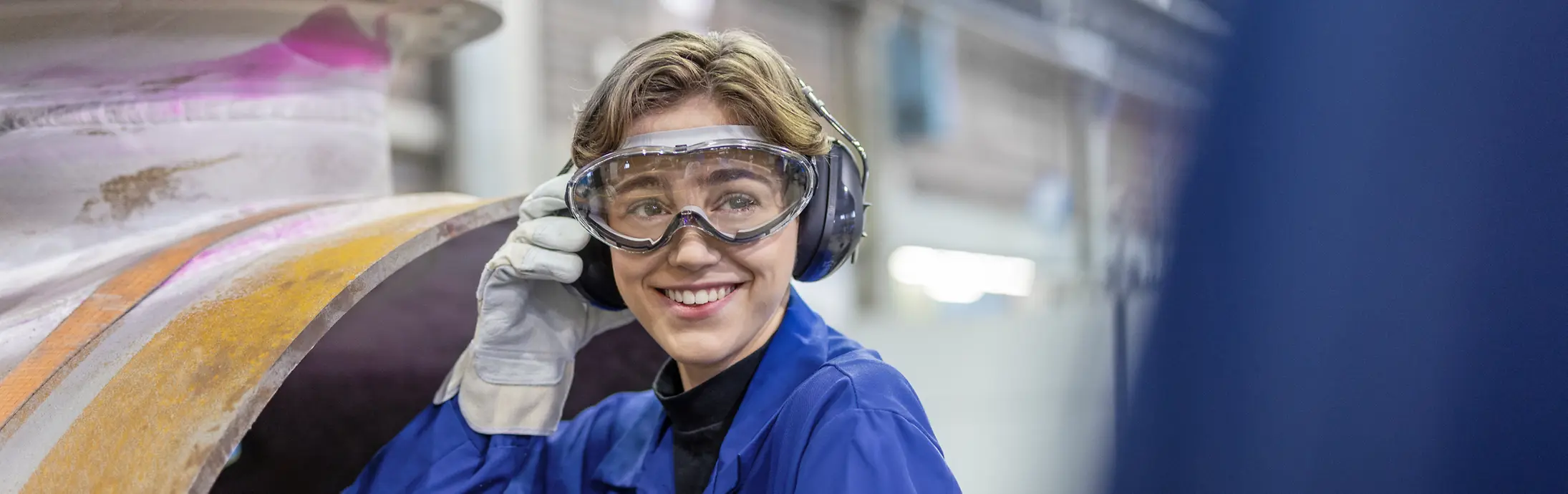 Eine junge Mitarbeiterin mit Schutzbrille und blauer Arbeitskleidung an einem Produktionsstandort