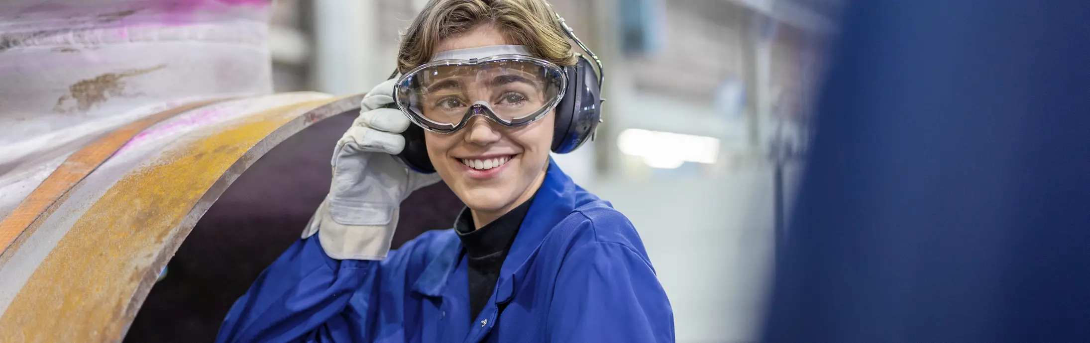 Eine junge Mitarbeiterin mit Schutzbrille und blauer Arbeitskleidung an einem Produktionsstandort