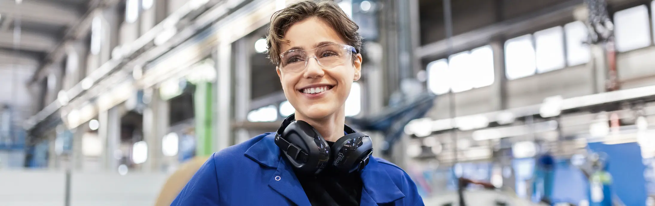 Eine junge Mitarbeiterin mit Schutzbrille und blauer Arbeitskleidung an einem Produktionsstandort