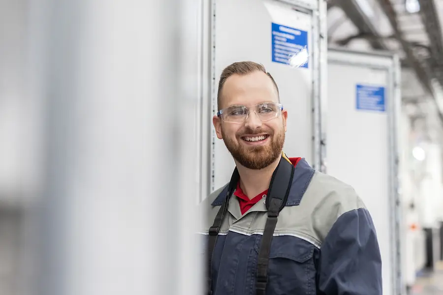 Junge Mitarbeitende mit Schutzbrille und blauer Arbeitskleidung an einem Produktionsstandort
