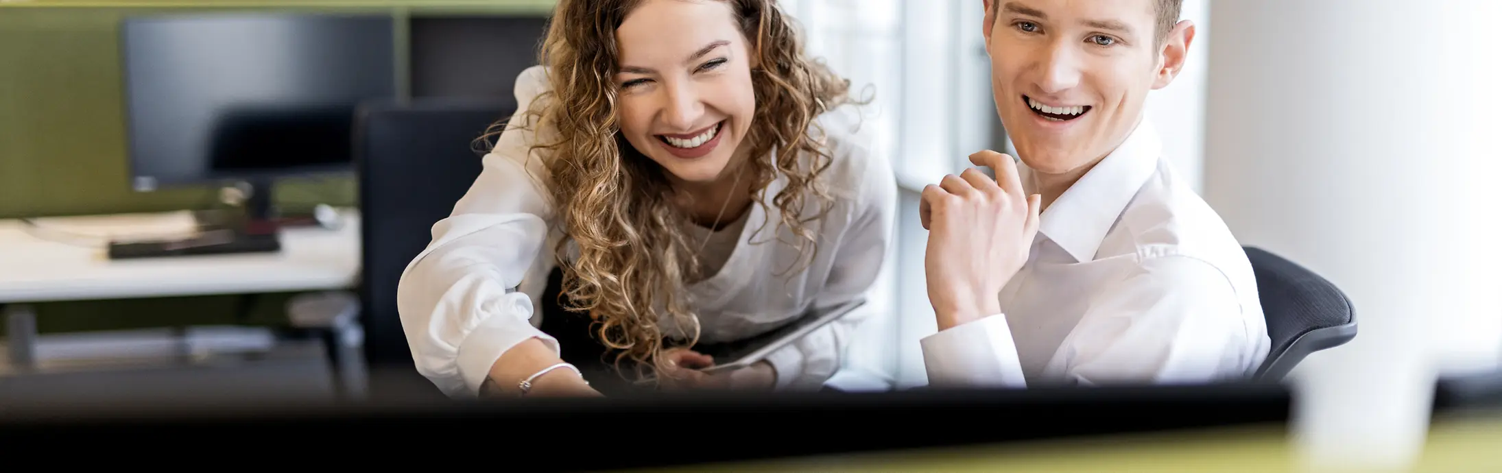 Zwei junge Mitarbeitende sitzen im Büro und schauen lächelnd auf einen Bildschirm