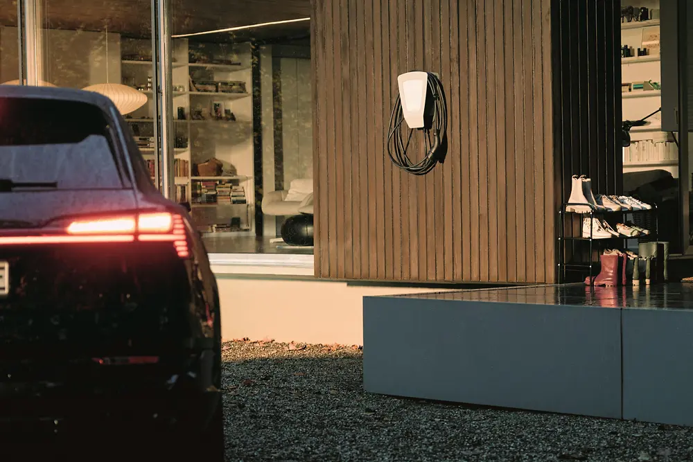 Dark electric car parked on a gravel driveway charging via a wall-mounted charging unit on a modern house with vertical wooden cladding. Warm interior lighting glows through large windows, showing shelves and furniture inside, while fallen leaves cover the ground in the foreground.