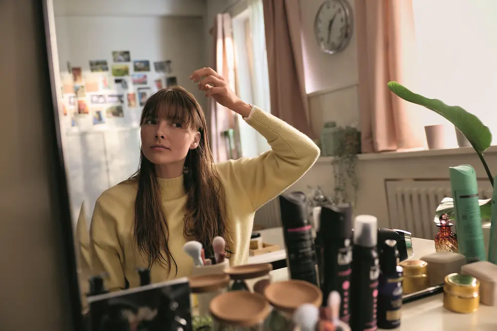 Person standing at a vanity table looking into a mirror while adjusting hair with one hand. The surface in front is filled with various beauty and haircare products, including bottles and brushes. Natural light from a window illuminates the room, with a plant, wall photos, and a clock visible in the background.