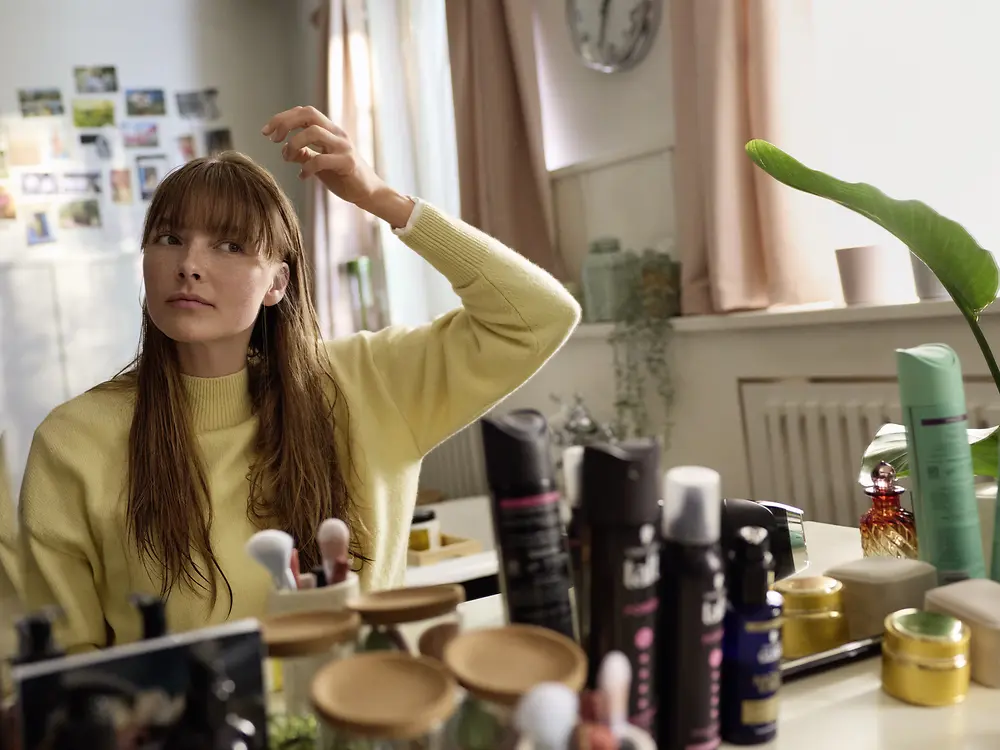 Eine junge Frau sitzt vor einem Spiegel und stylt ihr nasses Haar. Auf der Ablage stehen verschiedene Haarpflege- und Stylingprodukte.