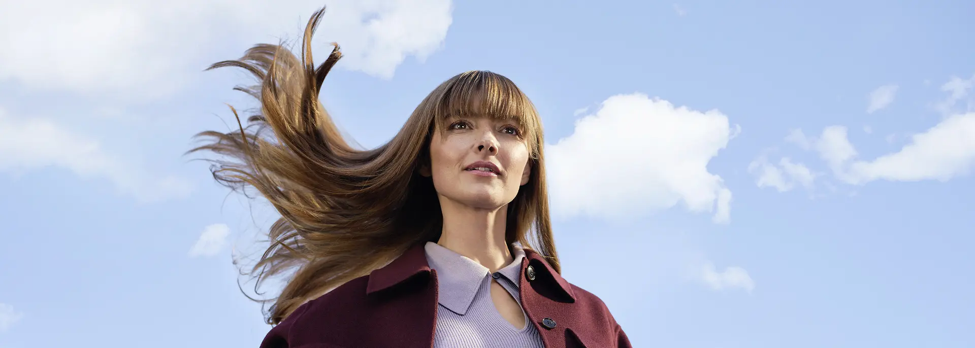 Junge Frau mit langen, im Wind wehenden braunen Haaren vor blauem Himmel und weißen Wolken.