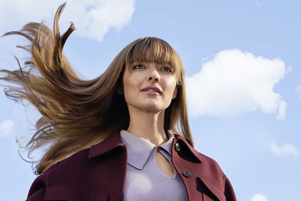 Junge Frau mit langen, im Wind wehenden braunen Haaren vor blauem Himmel und weißen Wolken.