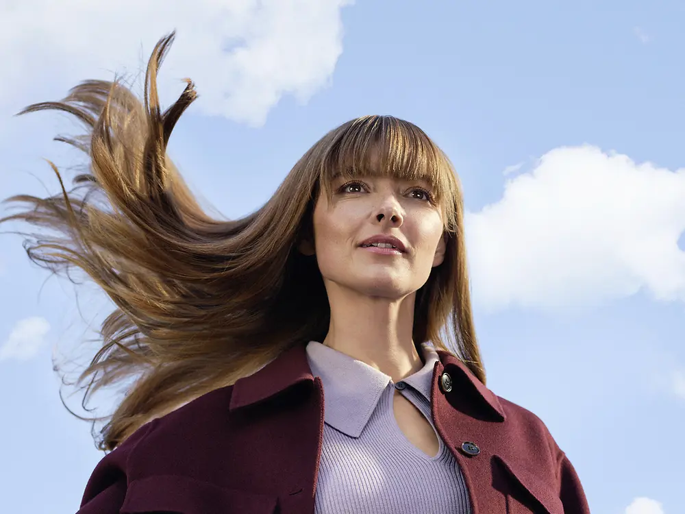Junge Frau mit langen, im Wind wehenden braunen Haaren vor blauem Himmel und weißen Wolken.