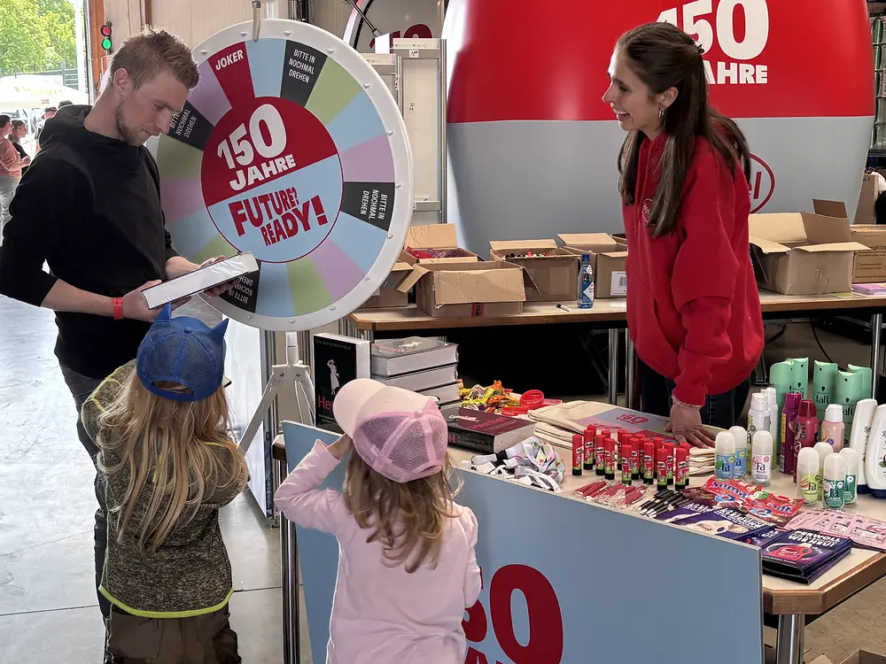 Ein Mitarbeiter steht mit seinen zwei Kindern vor dem Jubiläumsglücksrad. 