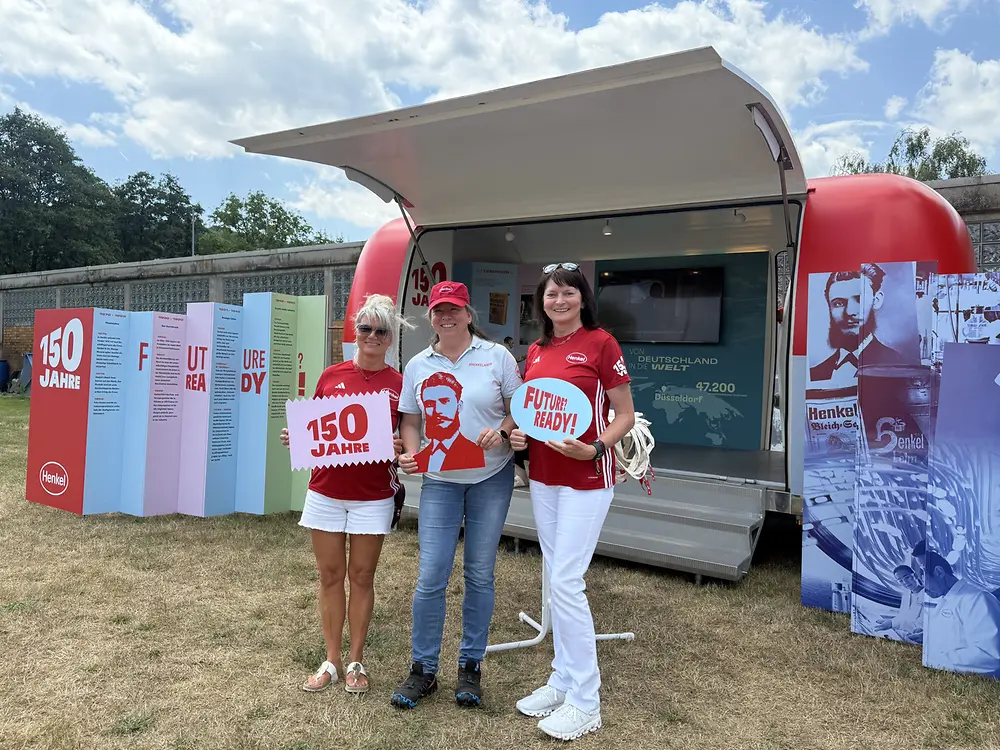 Drei Mitarbeitende stehen mit Postern vor dem Henkel-Jubiläumsmobil. 