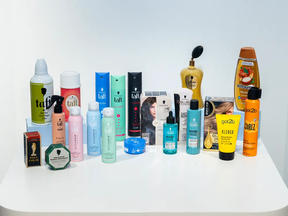 Historische und aktuelle Haarprodukte von Henkel, darunter Haarpflege- und Stylingprodukte in unterschiedlichen Verpackungsdesigns, nebeneinander präsentiert.
