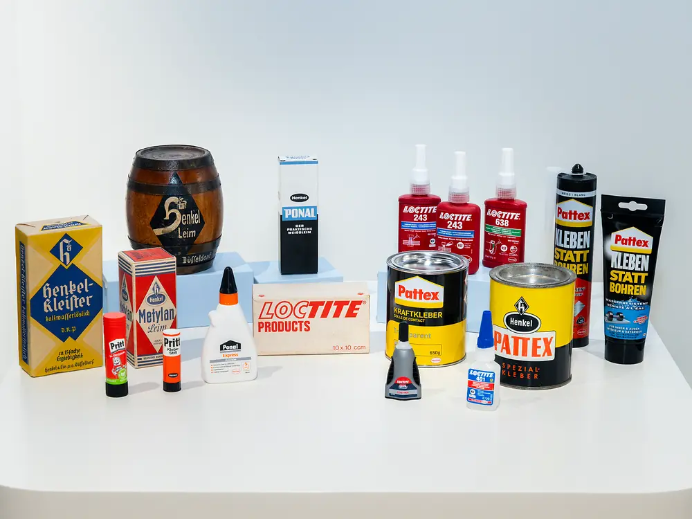 Historische und aktuelle Klebstoffprodukte von Henkel auf einem Podest präsentiert. Zu sehen sind Produktverpackungen der Marken Pritt, Ponal, Loctite, Pattex und Henkel aus unterschiedlichen Zeitperioden.