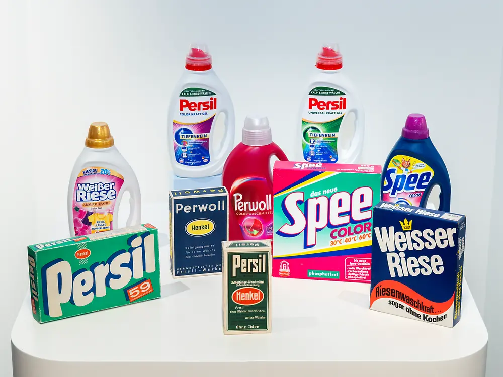 Historische und aktuelle Waschmittelprodukte von Henkel auf einem Podest präsentiert. Zu sehen sind Verpackungen und Flaschen der Marken Persil, Spee, Perwoll und Weißer Riese in unterschiedlichen Designs.