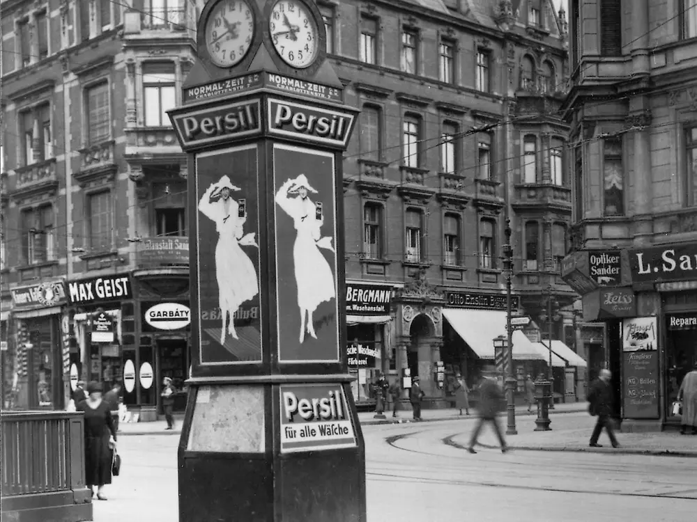 Historische Persil-Werbeuhr auf einem Platz in einer Innenstadt, umgeben von Gebäuden und Passanten.