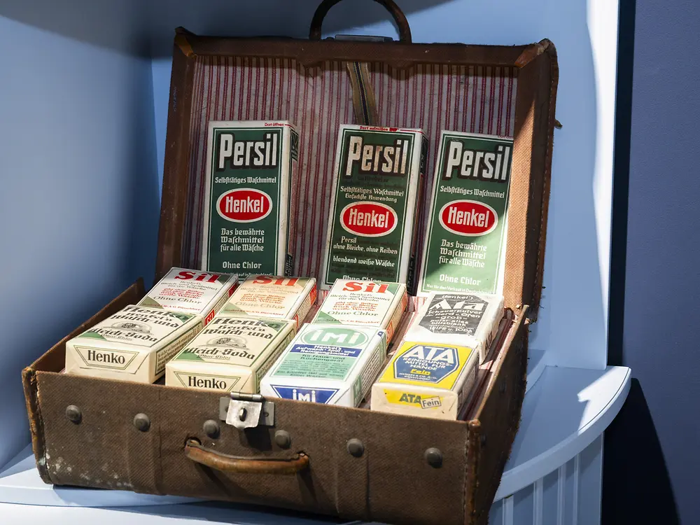 Historischer Verkaufskoffer mit verschiedenen Produktverpackungen von Persil und Henkel. Die Verpackungen sind aufrecht und liegend in einem geöffneten Koffer präsentiert.