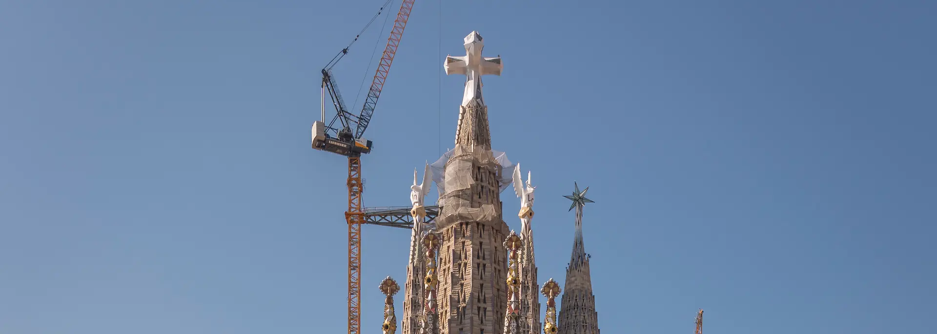 Frontalansicht der Sagrada Família in Barcelona. Im Vordergrund sind die umliegenden Stadtgebäude zu sehen, hinter der Basilika ragen mehrere Baukräne empor. Darüber spannt sich ein klarer blauer Himmel.